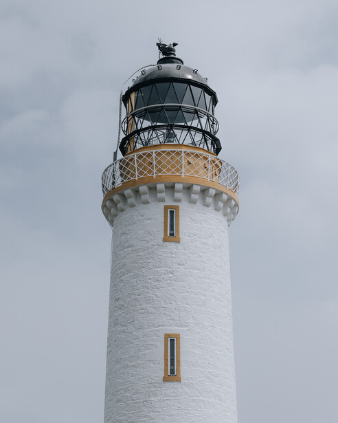 White lighthouse tower with brass gallery railings and lantern room against overcast sky.