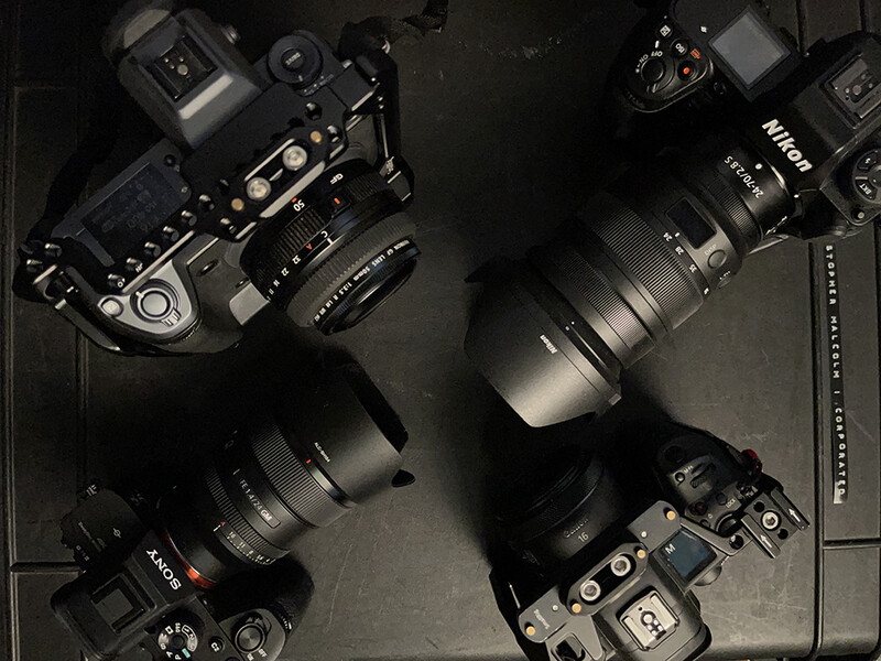 Four mirrorless cameras with various lenses arranged on a dark surface, shot from above.
