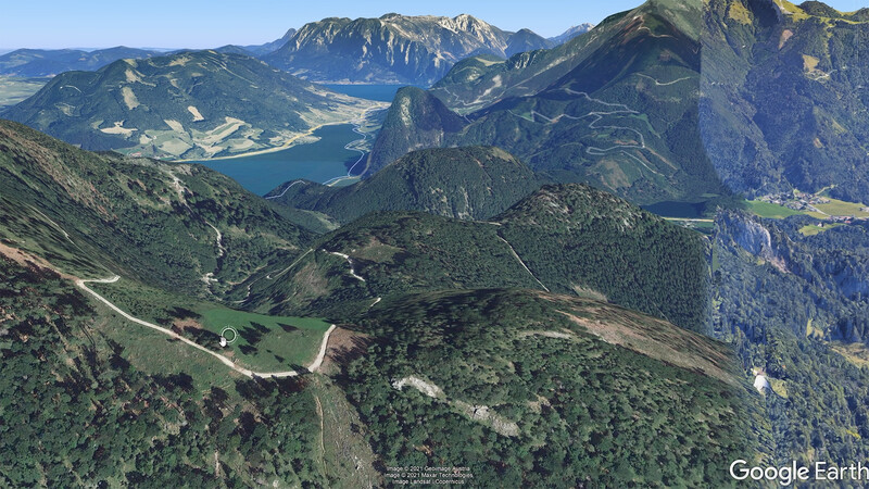 Aerial view of a mountainous Alpine valley with a winding road, forested slopes, and a turquoise lake nestled between peaks.