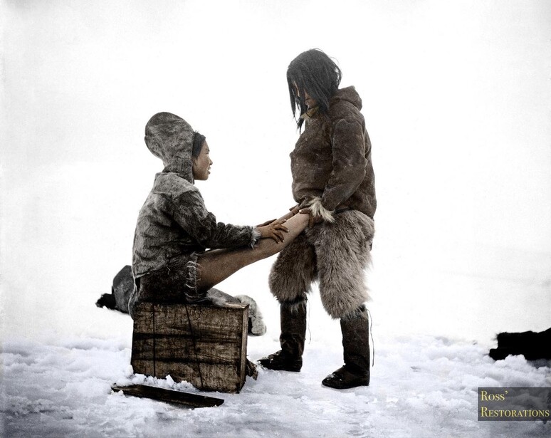 Two figures in heavy fur clothing kneeling face-to-face on snow with a wooden crate nearby.