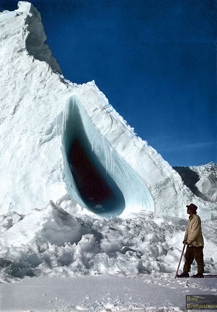 Massive crevasse with deep blue interior cut through white glacier, person standing nearby for scale.