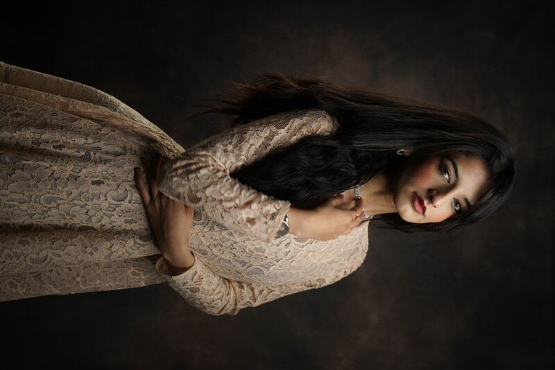 Woman with long dark hair wearing a cream lace dress against a dark textured background.