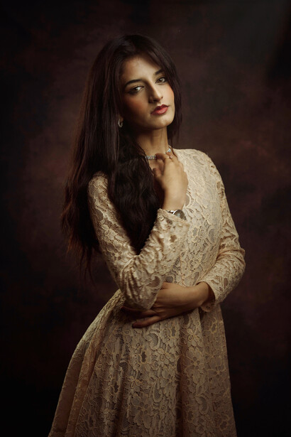 Woman with long dark hair wearing a cream-colored lace dress against a dark textured background.