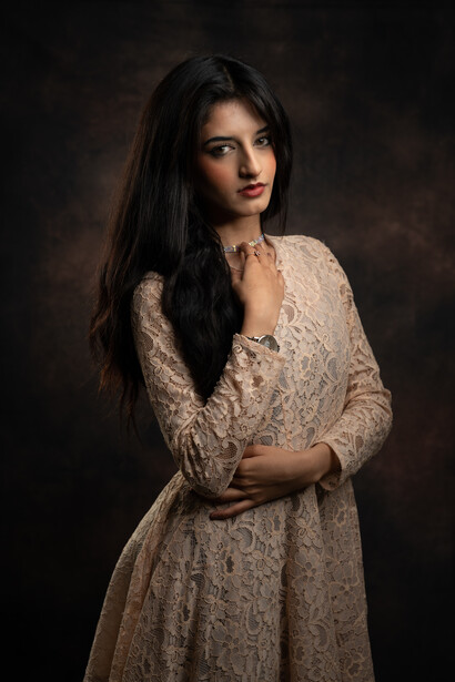 Woman with long dark hair wearing a beige lace dress against a dark brown background.