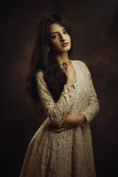 Woman with long dark hair wearing an embellished cream dress against a dark brown background.