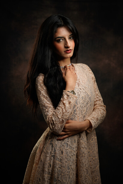 Woman with long dark hair wearing a cream lace dress against a dark background.