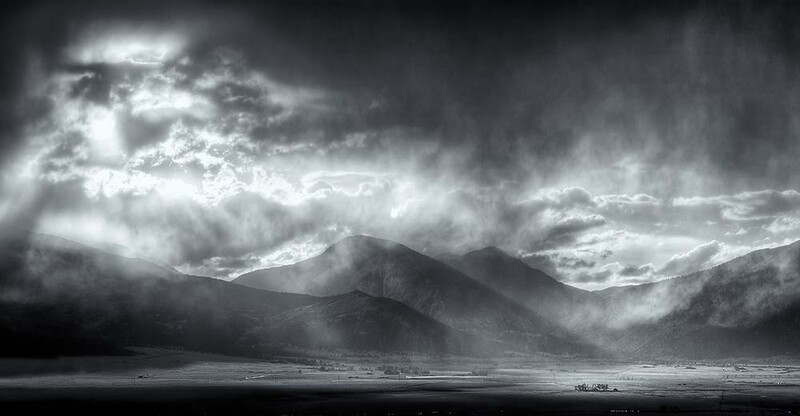 Dramatic black and white landscape with mountains silhouetted beneath turbulent storm clouds and breaking light rays.
