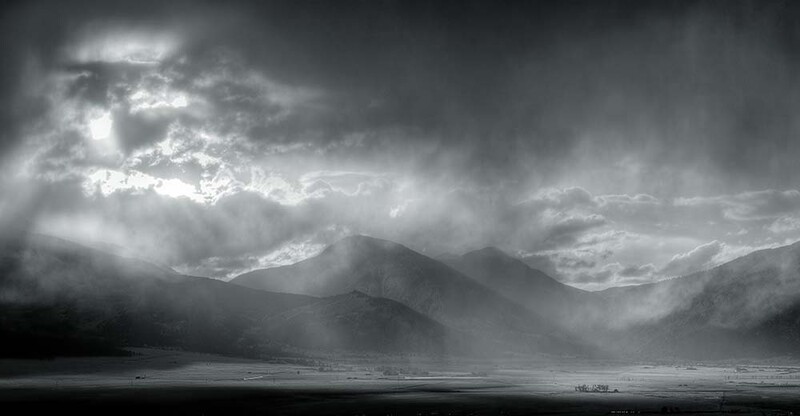 Dramatic black and white landscape with layered mountains beneath stormy clouds and crepuscular rays.