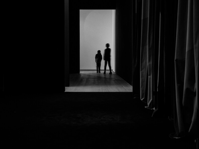 Two silhouetted figures standing in a bright doorway viewed from a dark interior space with draped curtains.