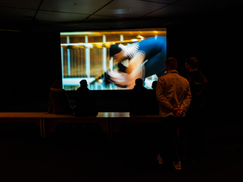Silhouetted audience members watching a large projected image of a person in blue clothing against a bright interior background.