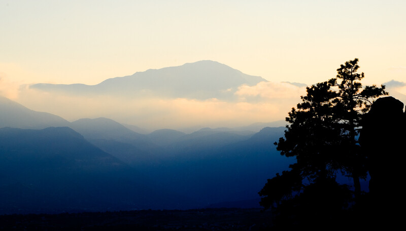 Silhouetted tree in foreground with layered mountain ridges receding into misty distance at dusk.
