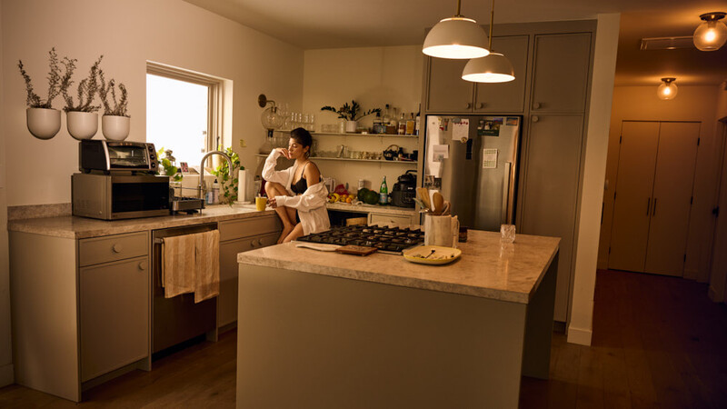 Woman cooking at a gas cooktop in a warm-toned kitchen with a large island and pendant lighting.