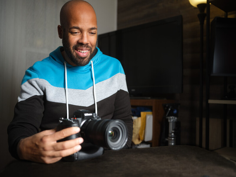 Photographer holding a DSLR camera in a studio setting with professional lighting equipment visible in the background.