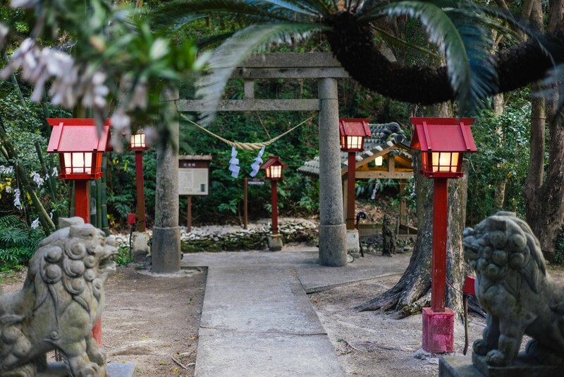 Japanese temple gateway with red lantern posts, stone lion statues, and lush green foliage.