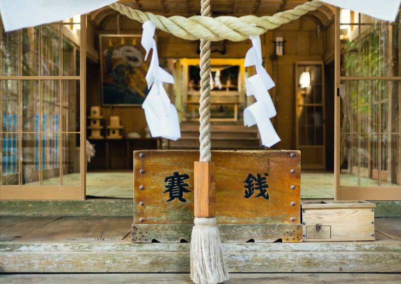 Wooden shrine entrance with rope, white paper strips, and metal plaque with Japanese characters.