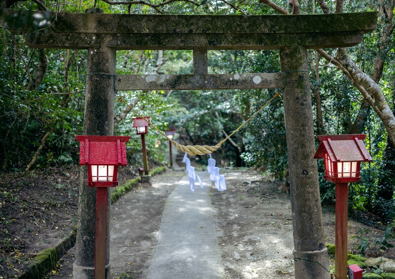 Stone torii gate frames a pathway where two figures in white walk toward a shrine entrance surrounded by lush greenery.