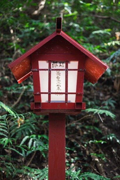 Red wooden birdhouse with white trim and peaked roof mounted on a red post against green foliage.