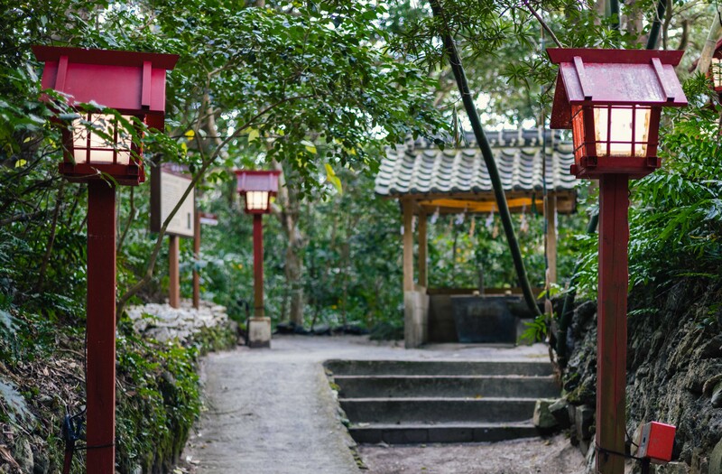 Red wooden lantern gates frame a stone pathway through lush greenery at a traditional East Asian temple or garden entrance.