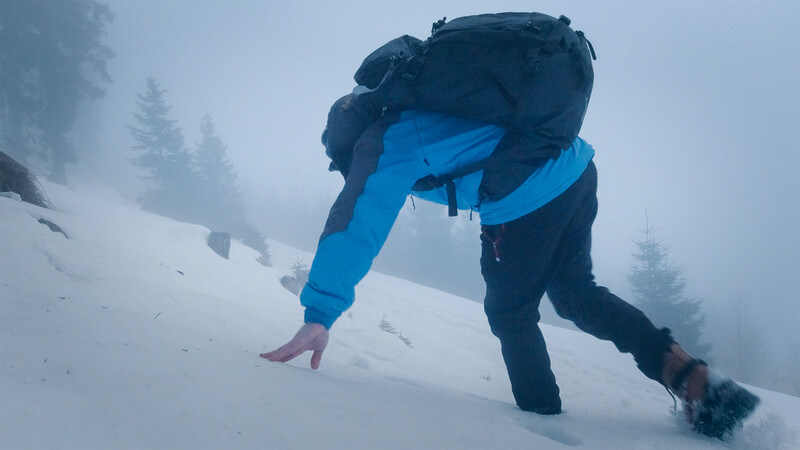 Hiker in blue jacket bending forward on snowy slope in heavy fog with evergreen trees.