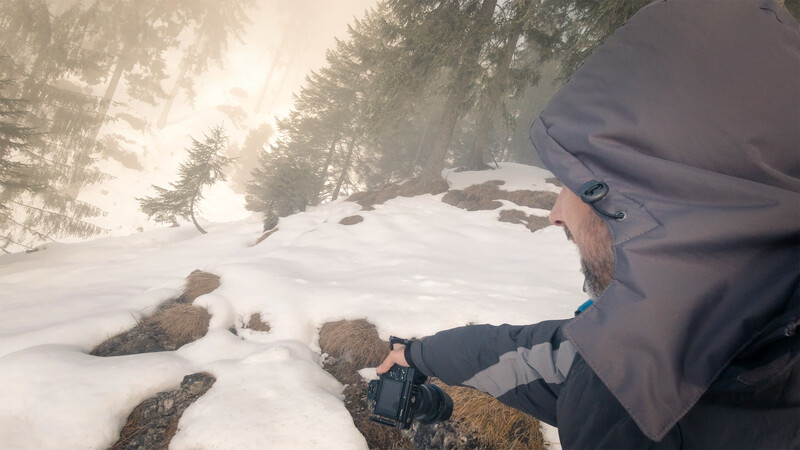 Photographer holding a camera on a snowy mountain slope surrounded by evergreen trees and rocky outcrops.