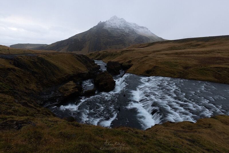 Snow-capped mountain peak rising above a glacial stream cutting through barren moorland.