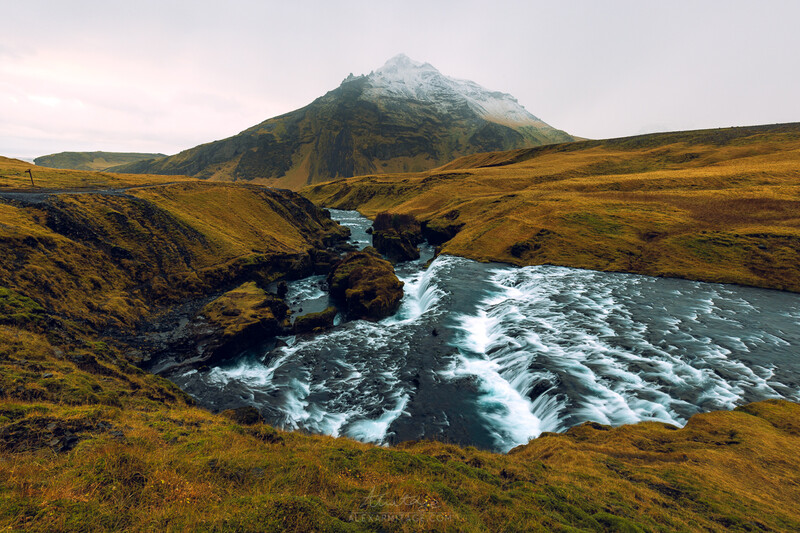 Snow-capped mountain rising above a glacial river cutting through golden tundra landscape.