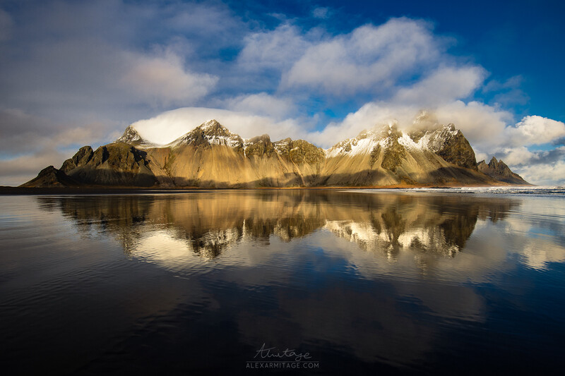 Snow-capped mountain peaks reflected in still dark water under blue sky with white clouds.