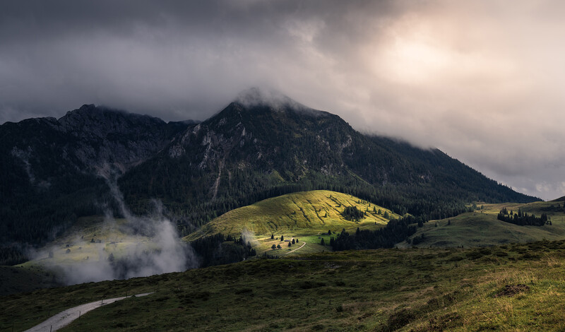 Mountain landscape with dramatic clouds, forested slopes, and a geothermal steam plume rising from a valley.