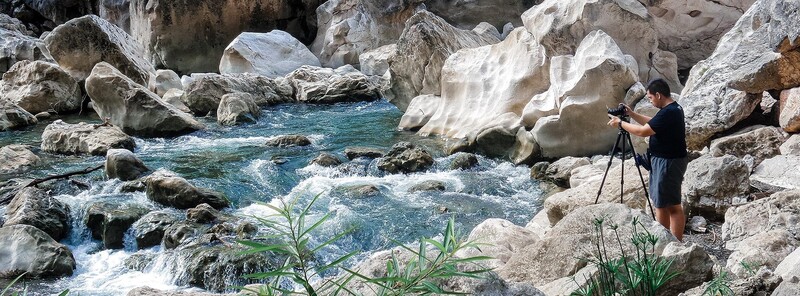 Photographer with tripod shooting a mountain stream surrounded by large boulders and turquoise water.