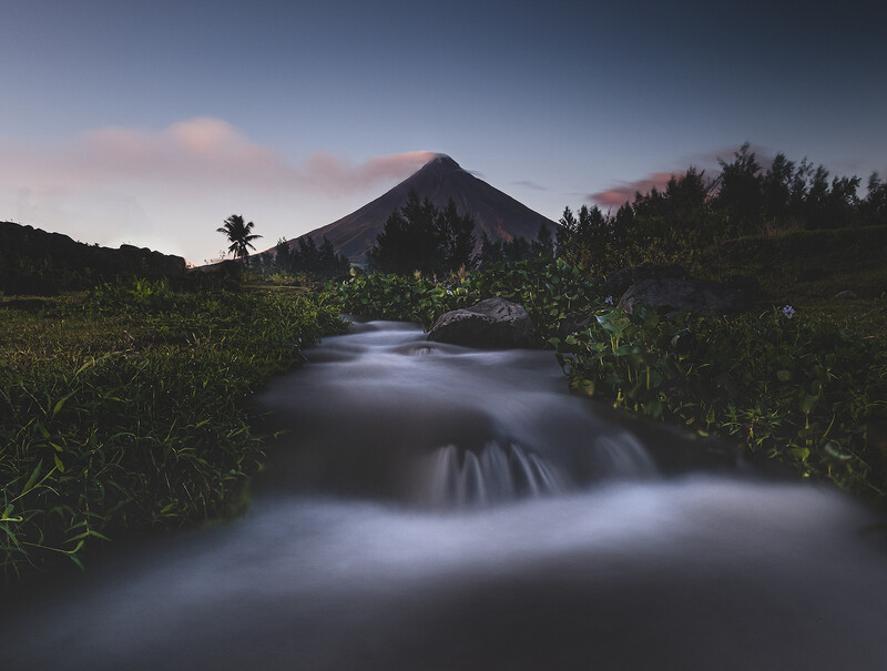 River with long exposure blur flowing toward a conical volcano at dusk.