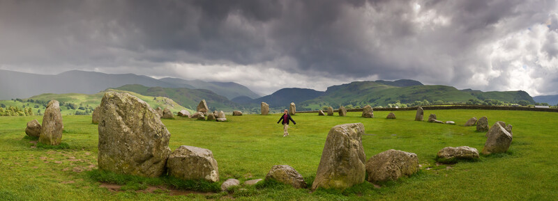 Ancient stone circle set in expansive green valley beneath dramatic cloudy sky and distant mountains.