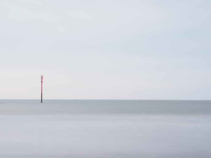 Single red navigation buoy standing alone in calm, misty water under overcast sky.