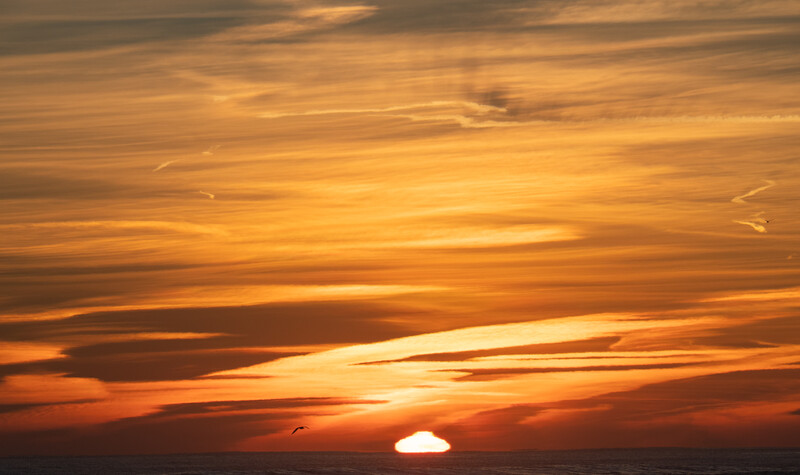 Sun setting below the horizon with layered orange and gold clouds stretching across the sky.