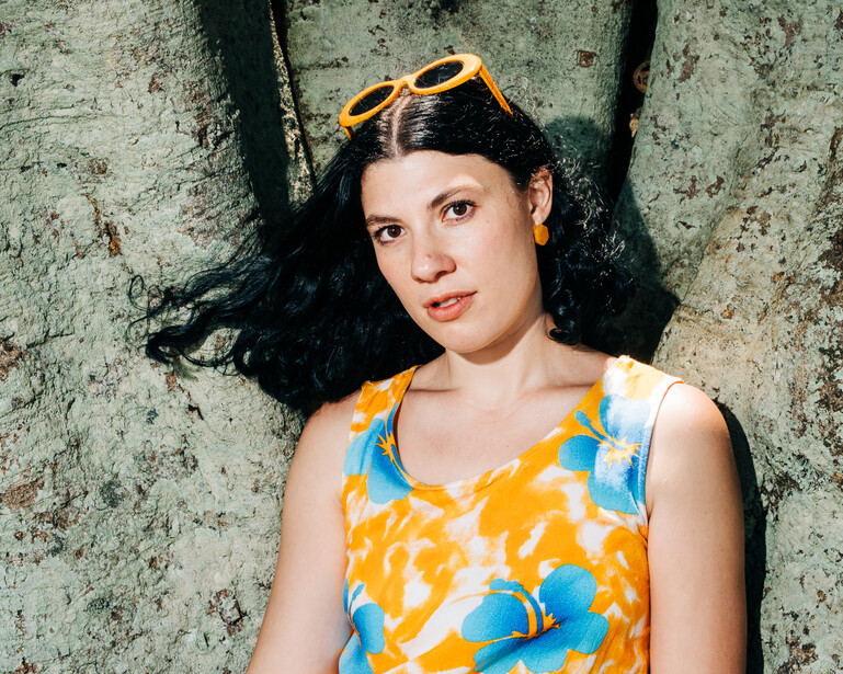Woman with long dark hair wearing yellow and blue patterned sleeveless top, sunglasses on head, posed against weathered concrete wall.