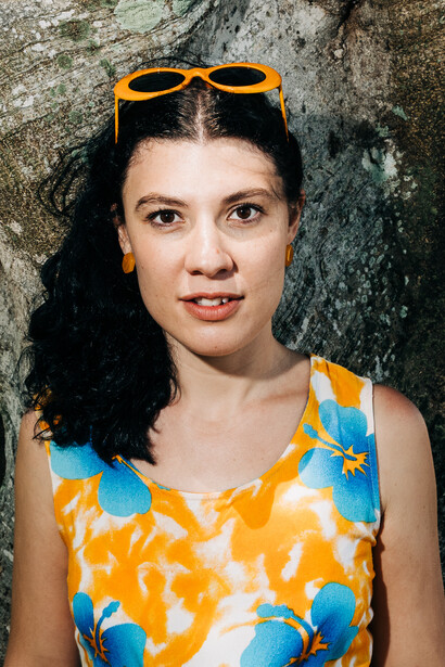 Woman with long dark hair wearing yellow and blue floral top, orange sunglasses pushed up on head, posed against weathered concrete wall.