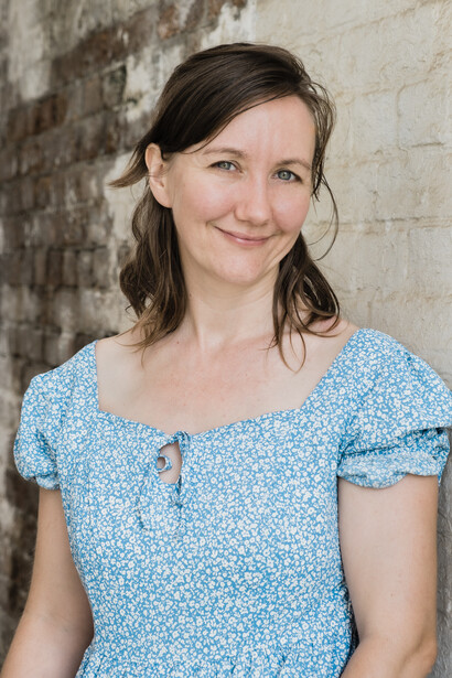 Woman with shoulder-length dark hair wearing a light blue floral short-sleeve top, smiling at camera against a weathered brick wall.
