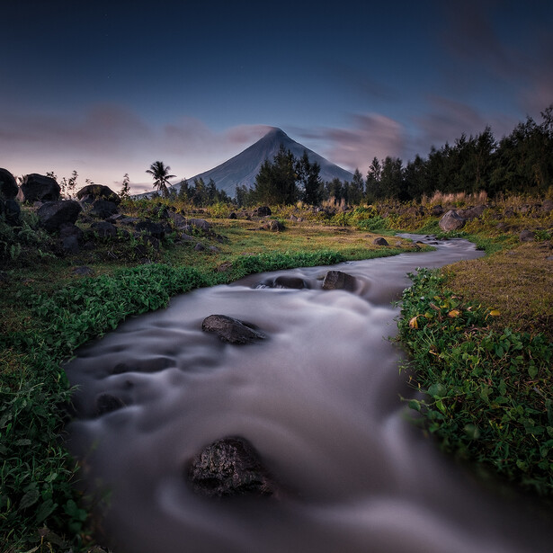 A cone-shaped volcano rises behind a flowing stream with long-exposure blur, surrounded by grassland and forested slopes at dusk.