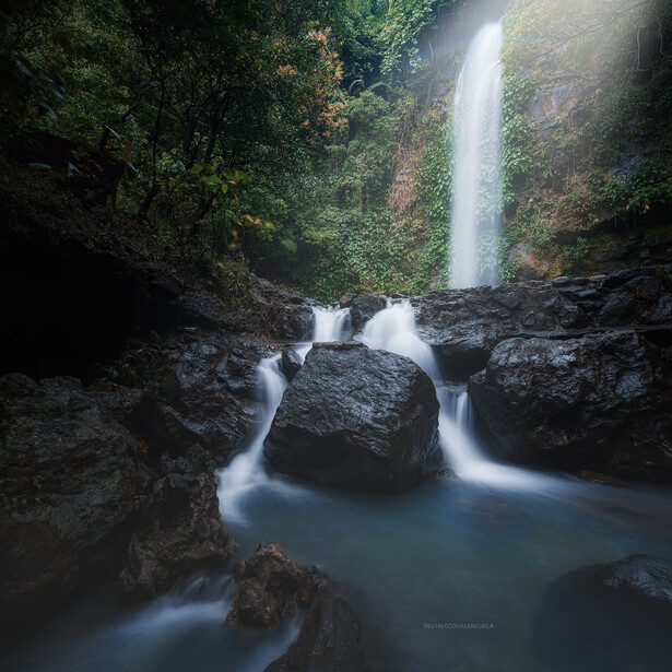 Waterfall cascading into a misty pool surrounded by lush green vegetation and dark rocks.