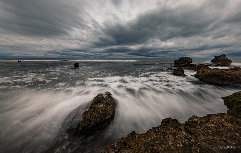 Waves crash over rocky outcrops on a shoreline beneath dramatic storm clouds.