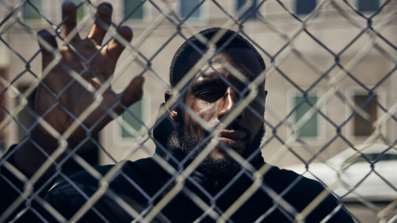 Person with dark hair and sunglasses behind chain-link fence, hand raised against the mesh.