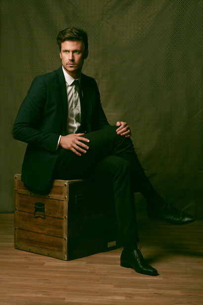 Man in dark green blazer and striped tie seated on wooden box against green backdrop.