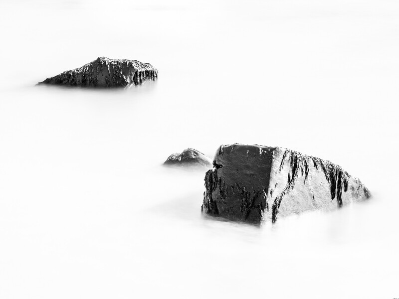 Two dark rock formations partially submerged in misty white water against a hazy sky.