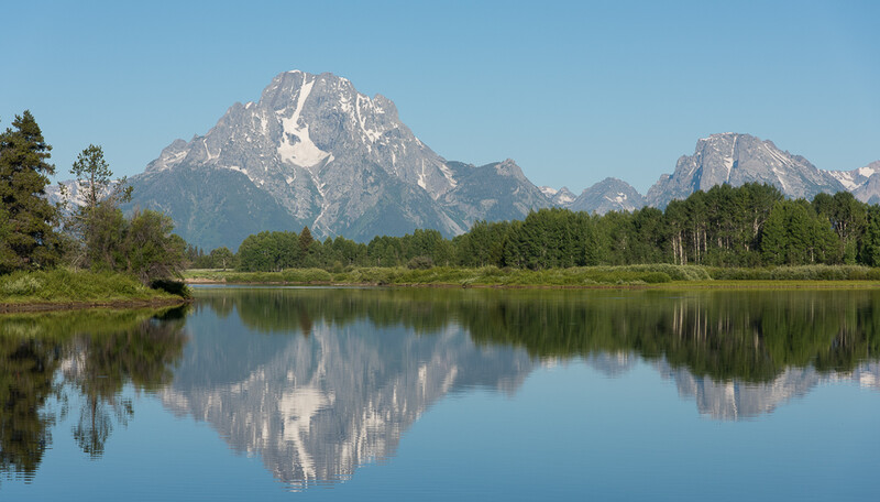 Snow-capped mountain peaks reflected in still river water, framed by evergreen forest.