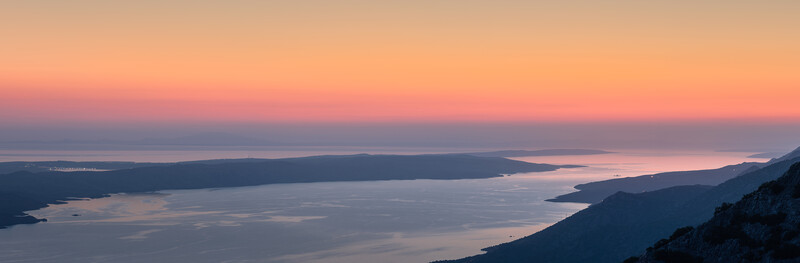 Mountainous coastline at sunrise with deep blue water and silhouetted peaks against a peachy-orange sky.