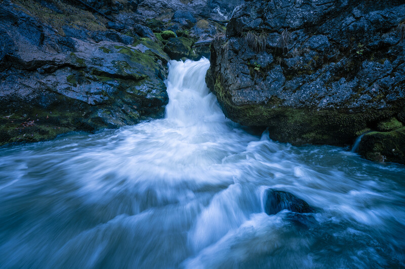 A cascading waterfall flows through moss-covered rocky canyon walls into a turquoise pool below.