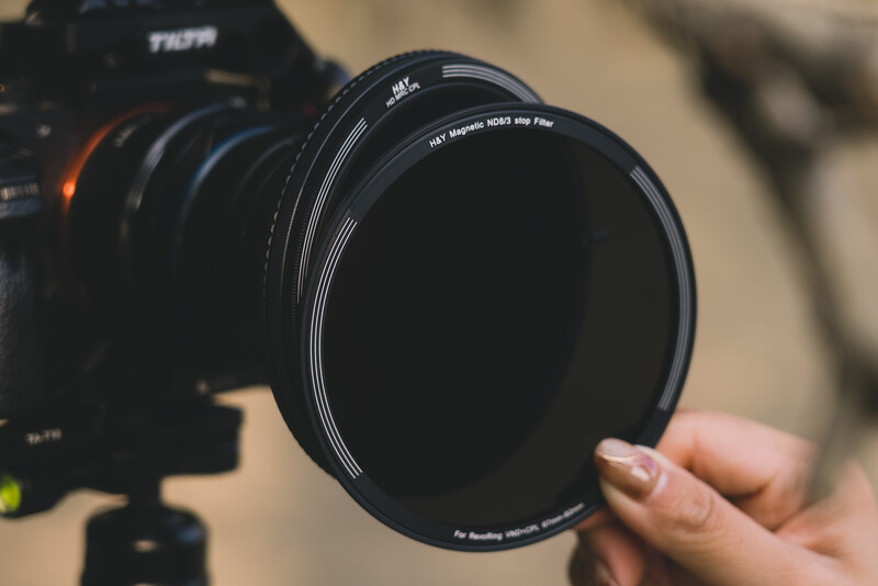 Photographer holding a circular neutral density filter in front of a camera mounted on a tripod.