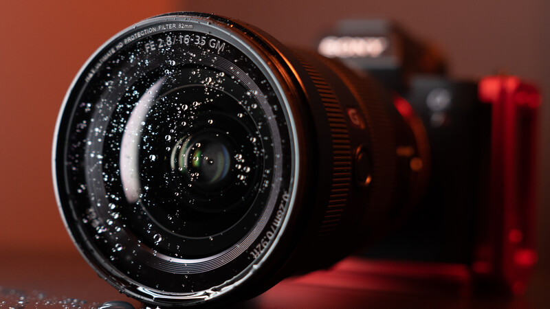 Wide-angle lens with water droplets on front element, mounted on camera body against warm background.