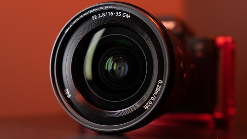 Sony FE 2.8/16-35 GM wide-angle zoom lens photographed against a warm orange background with camera body blurred behind.