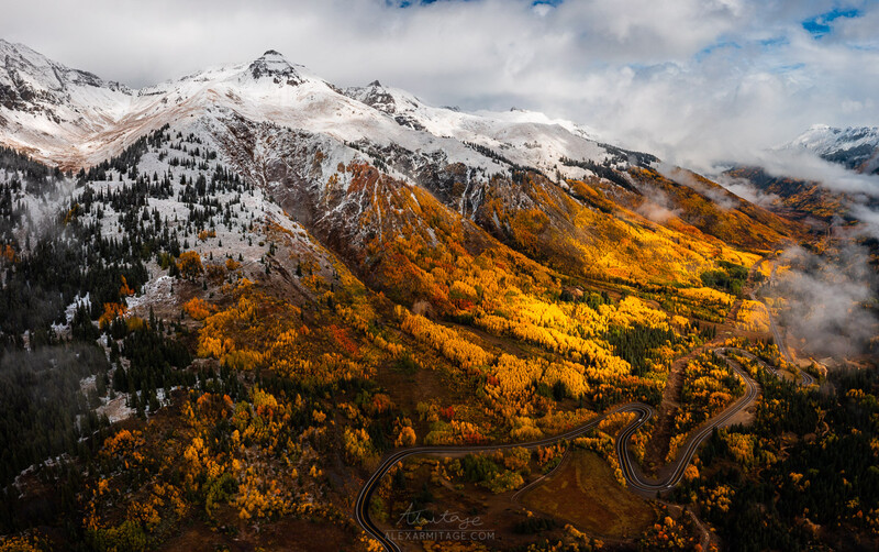Snow-capped mountains with golden larch trees in autumn valley below winding road.