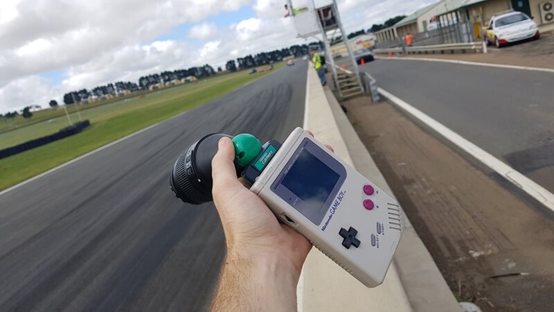 Hand holding a vintage Game Boy handheld console with a green cartridge at a racing circuit.